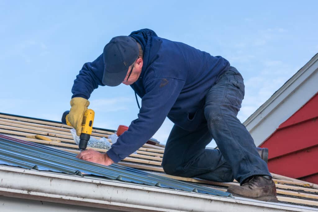 Couvreur avec perceuse jaune fixant des panneaux de toit en métal bleu sur une maison, en plein jour.