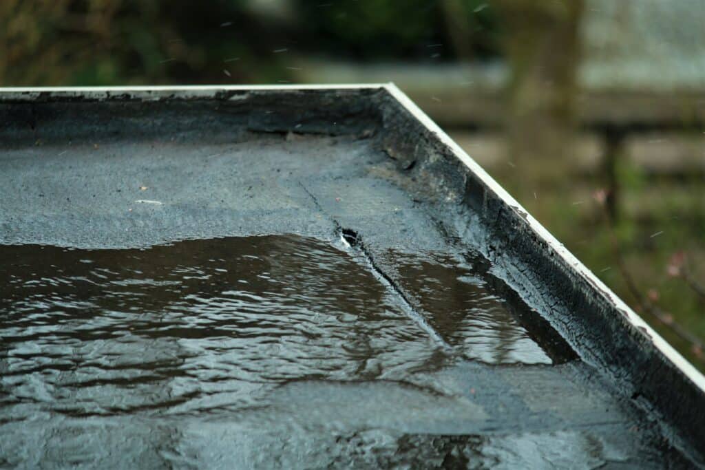 Surface sombre et fissurée d'un toit plat, recouverte d'eau stagnante et d'ondulations, sous une pluie fine. Arrière-plan flou vert.