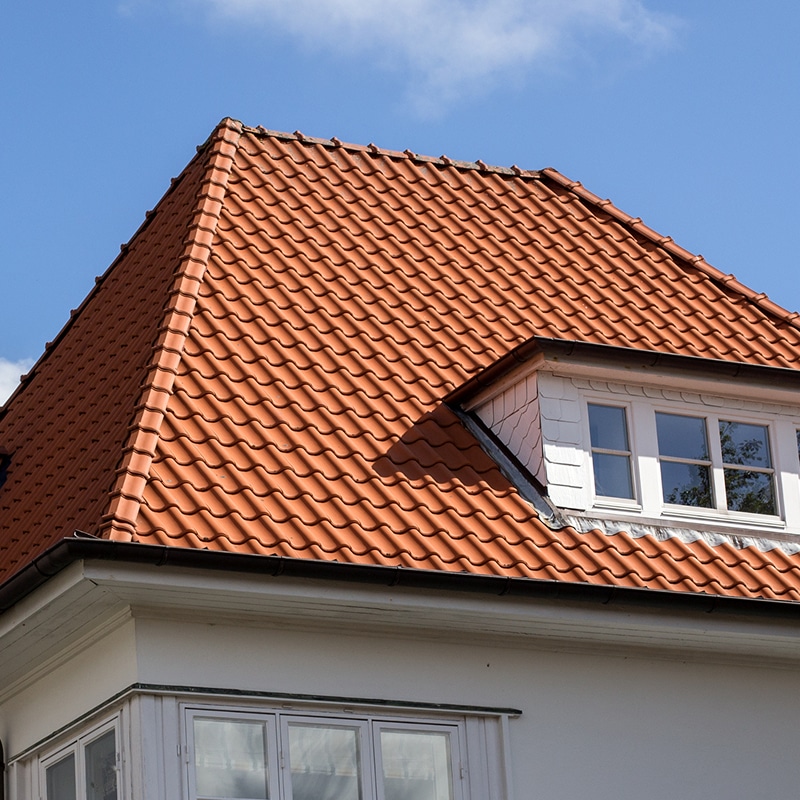 Toit en tuiles de terre cuite d'une maison blanche sous un ciel bleu. Une lucarne blanche avec des fenêtres est intégrée au toit.