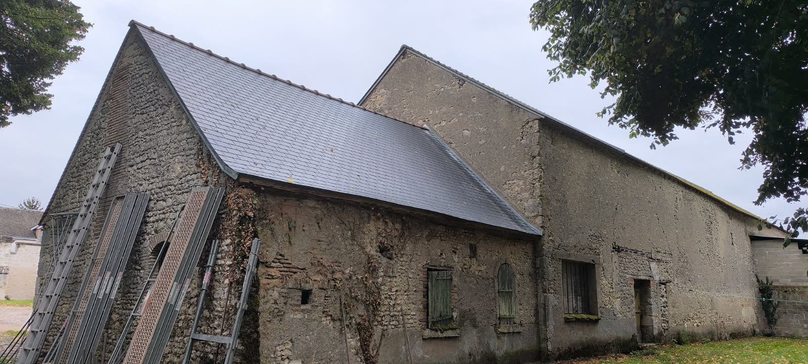 Ancienne grange en pierre avec toit en ardoise neuve, échafaudages et échelles contre le pignon lors de travaux.