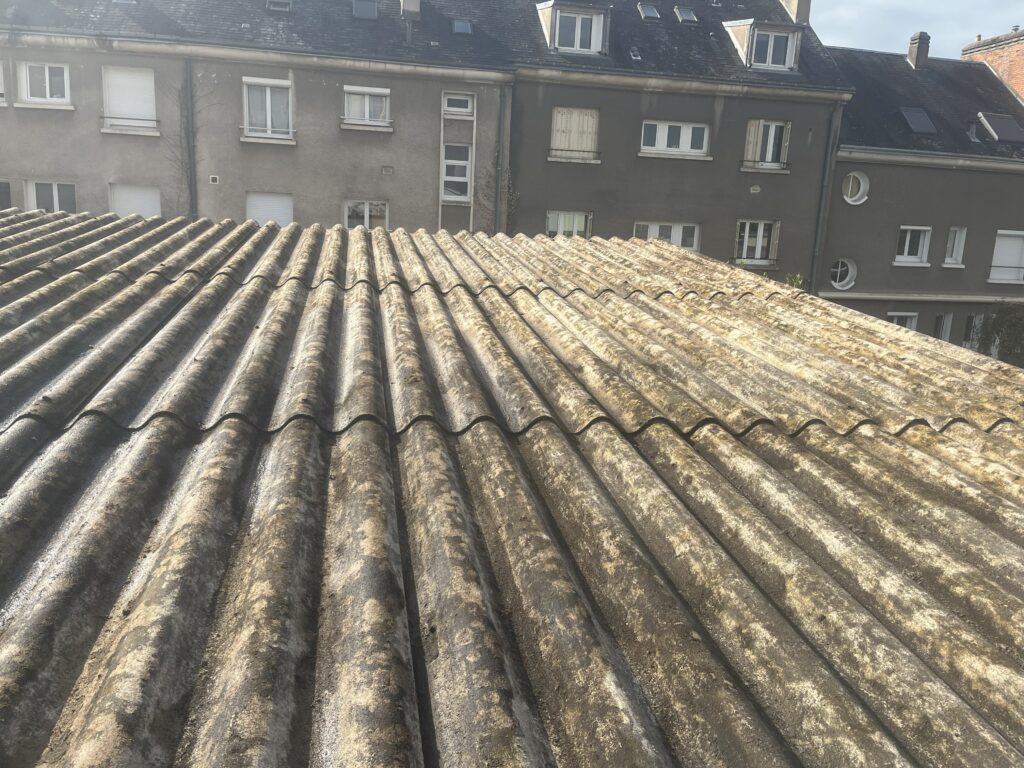Toit en plaques ondulées vieillies et jaunies par le temps, vue plongeante devant des façades d'immeubles gris avec volets.