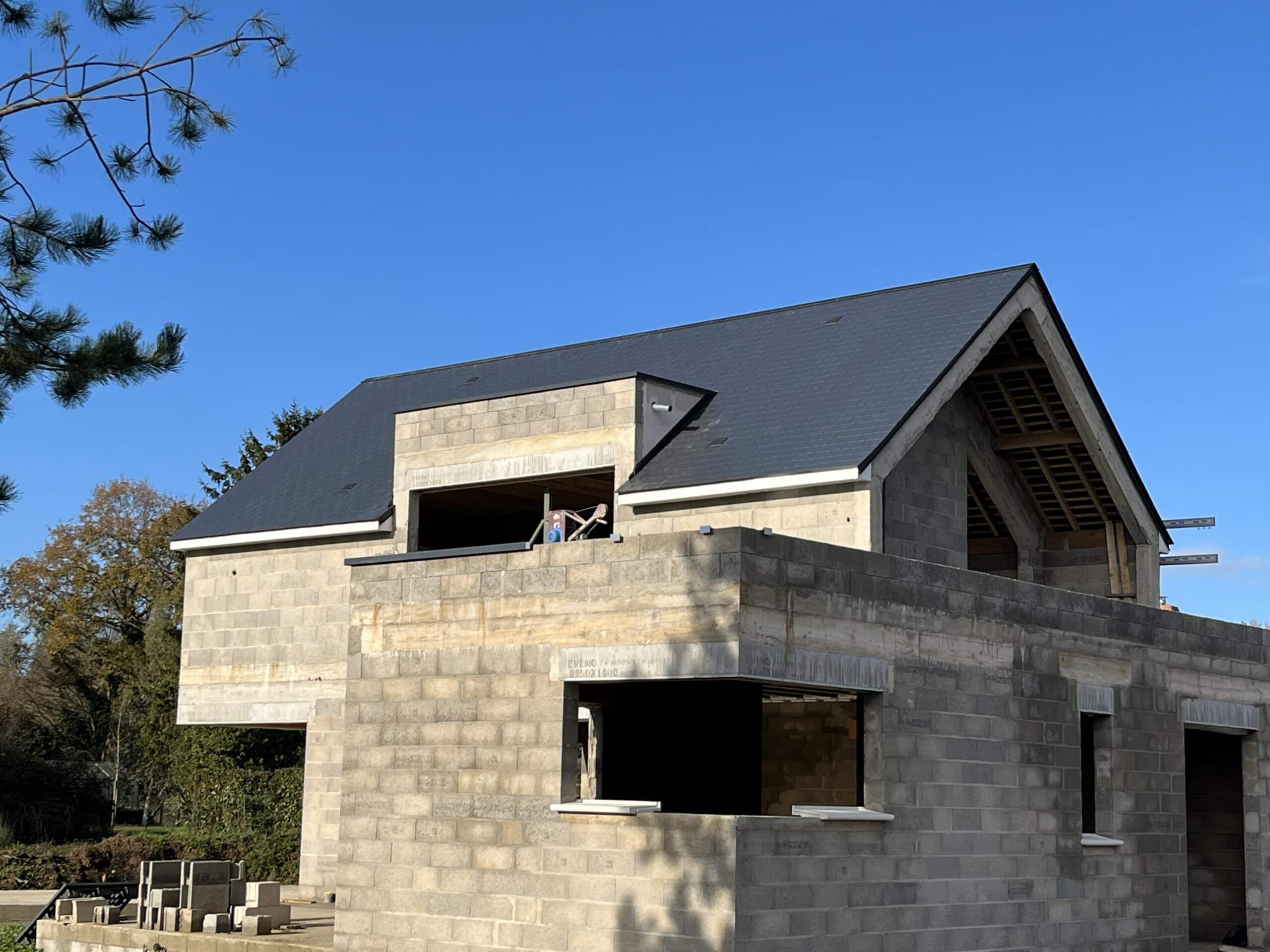 Construction maison en parpaings avec toiture en ardoise sombre. Un ouvrier est visible à une fenêtre de l'étage. Texte visible: EVENO 0950X1400.